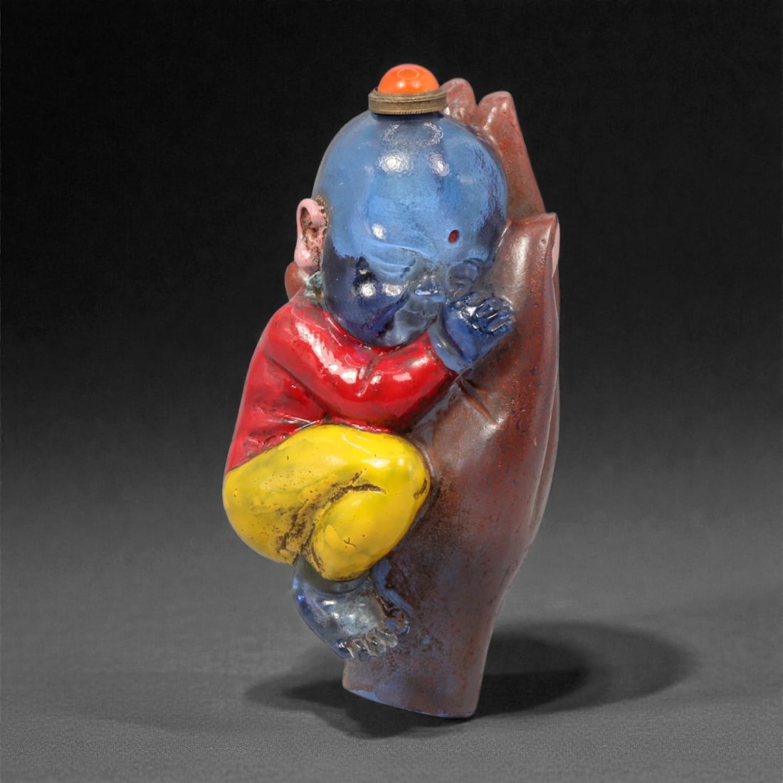 A Chinese Glass Figural Snuff Bottle, 20th Century, 59YC: Molded from translucent and opaque glass and finished with polychrome surface coloration, the snuff bottle is formed as a child shown asleep while leaning against an oversized hand. The figure is rend