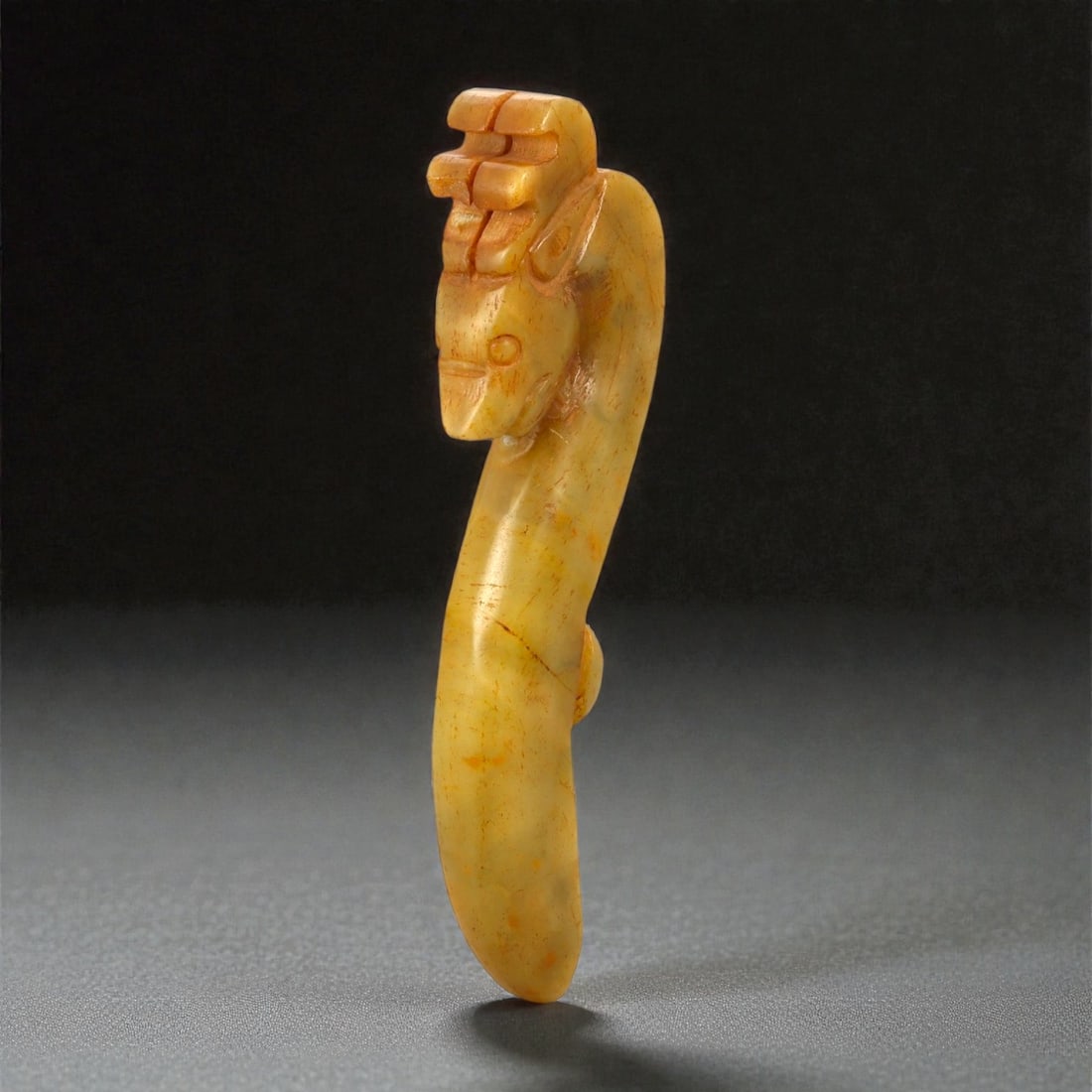 A Chinese Jade Belt Hook, 20th Century, 63FB: Carved from yellow-toned jade with areas of darker russet staining and natural inclusions, the belt hook is worked in a gently curved, elongated form. The terminal is carved as a stylized animal head,