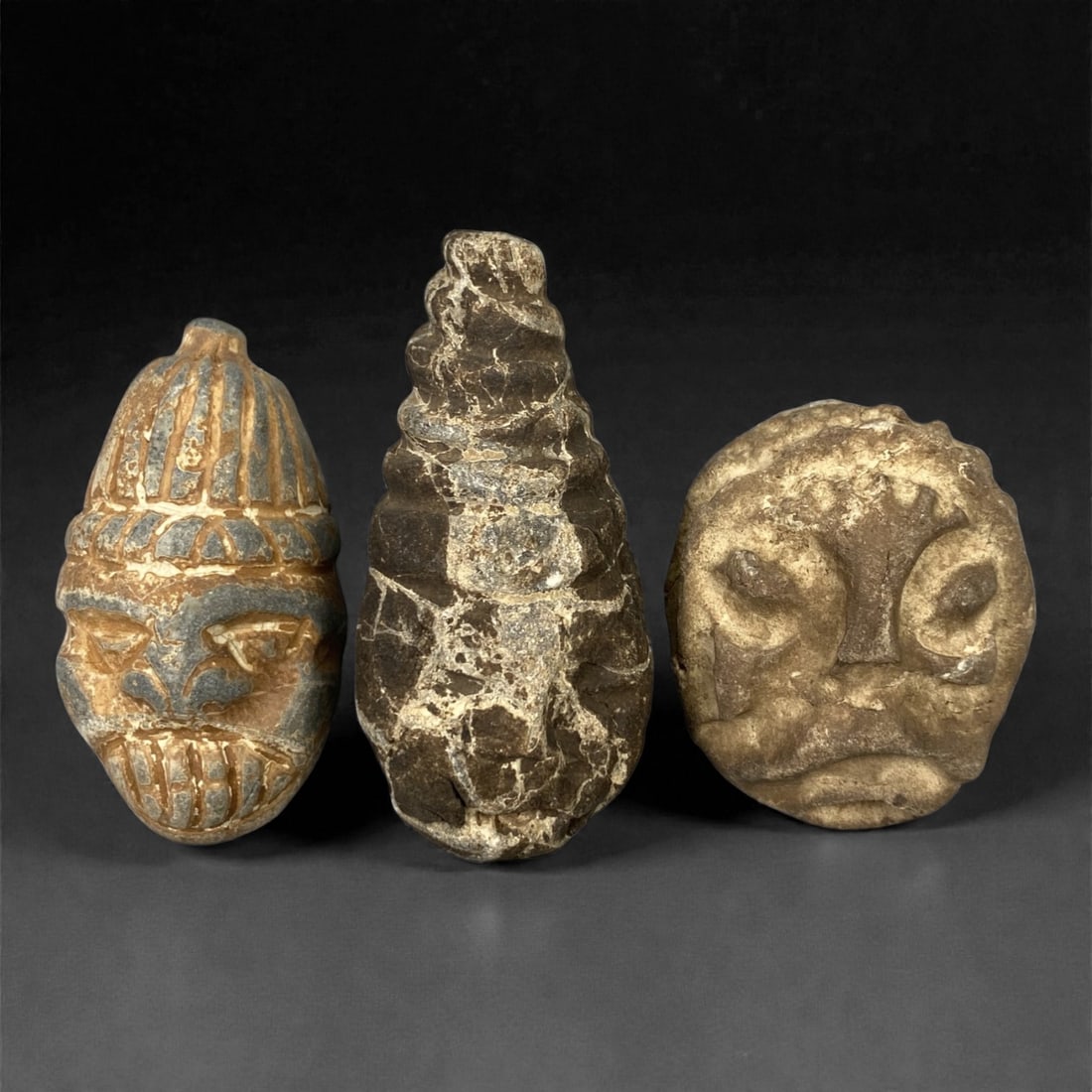 A Group of Three Ancient Stone Maskettes, 1st Millennium BCE, 76FM: Comprising three stone maskettes, each formed from light brown to grey stone with visible surface fissures and tonal variation. The leftmost maskette features a stylized face with a ribbed headdress,