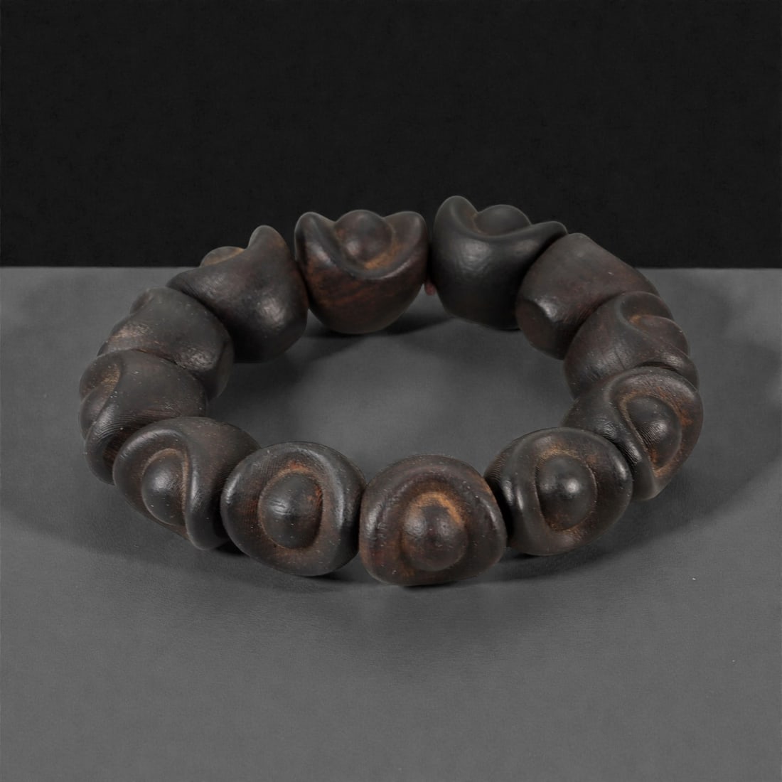 A Chinese Wood Bead Bracelet, 20th Century, 82FD: Composed of rounded beads carved from dark-toned wood, each bead modeled in a lobed, ingot-like form with softly rounded contours and a smooth, polished surface. The beads display natural tonal variat