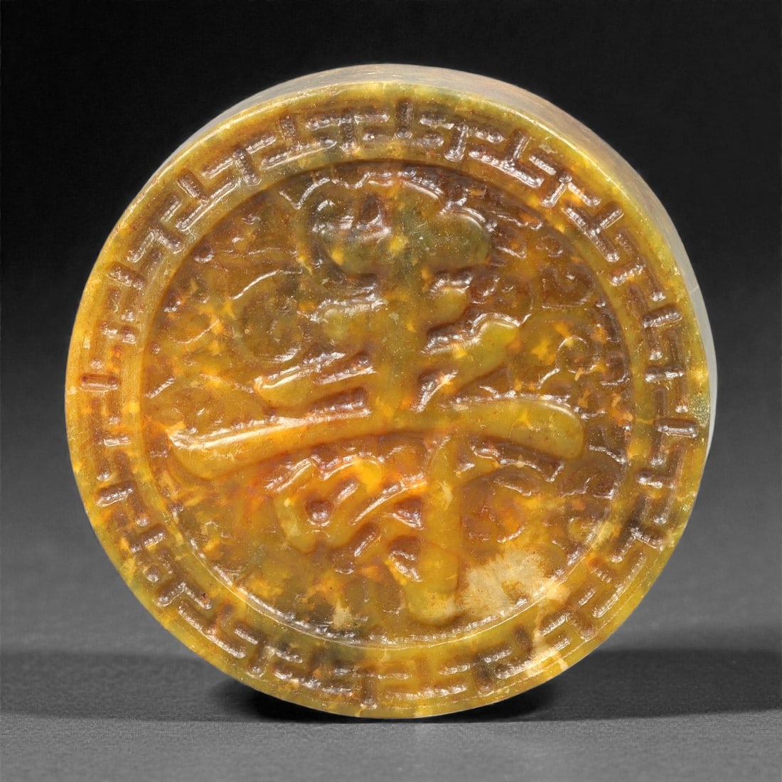 A Chinese Hetian Jade Shou Plaque, 20th Century, 47FH: Carved from mottled yellow-green Hetian jade with areas of darker veining and natural inclusions, the plaque is circular in form with a raised rim and shallow relief decoration. The central field is c