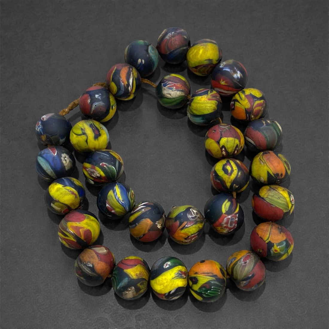 A West African Glass Mosaic Bead Necklace Strand, 19th-Early 20th Century, 287g, 16AA: Composed of spherical glass beads with a dark blue ground decorated with swirling, irregular patterns of yellow, red, and green, strung on a modern braided fiber cord. The multi-colored mosaic effect