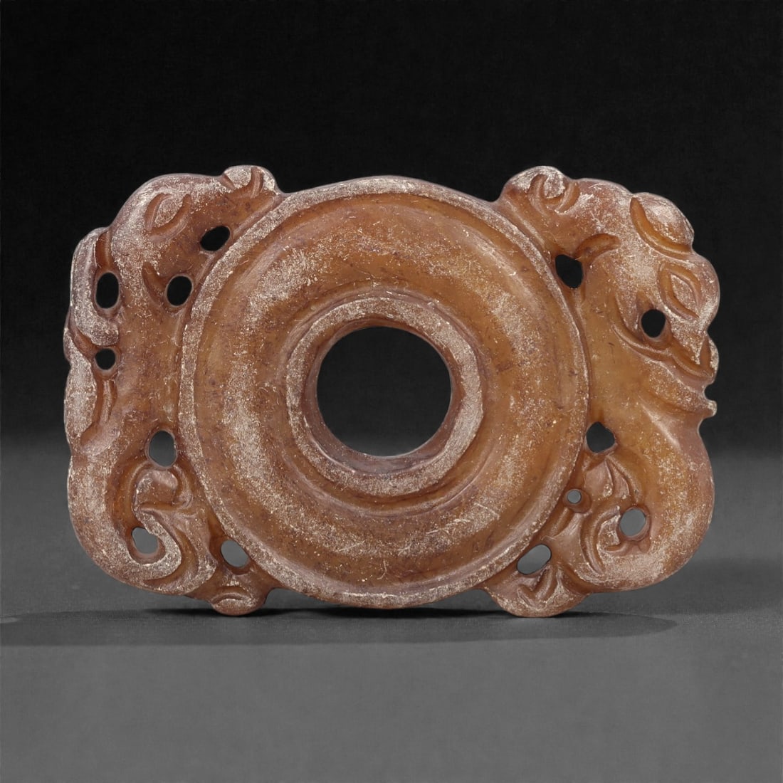 A Chinese Hetian Jade Openwork Plaque, 20th Century, 82FH: Carved from warm brown-toned Hetian nephrite jade with lighter translucent areas and natural surface mottling, the plaque is worked in an openwork format centered on a circular aperture. The surroundi