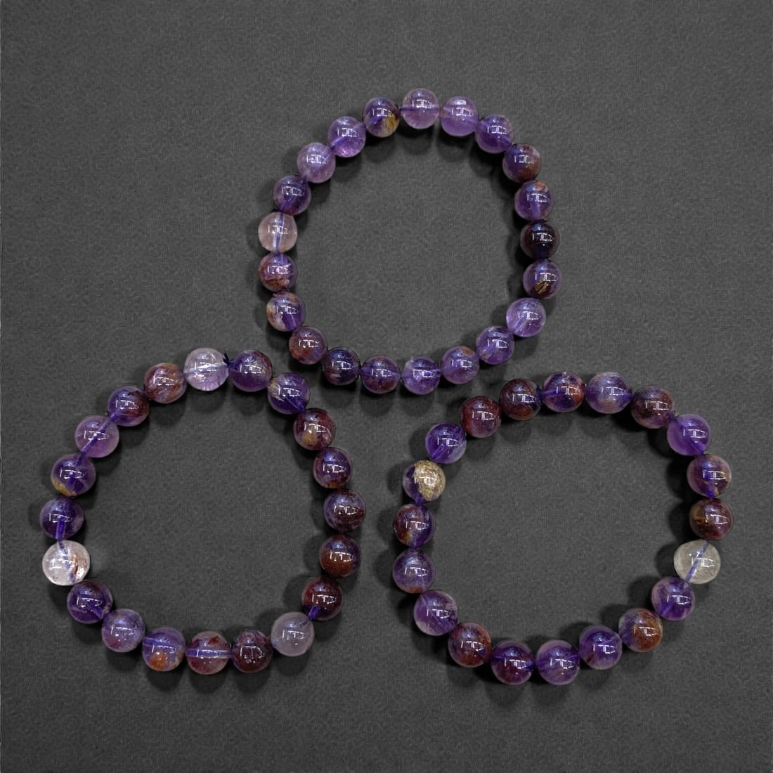 A Group of Three Super Seven Amethyst Beaded Bracelets, 77g, 76AA: Comprising three stretch bracelets composed of polished spherical Super Seven amethyst beads. The beads exhibit a range of hues, including deep violet, lavender, and russet, with natural internal incl
