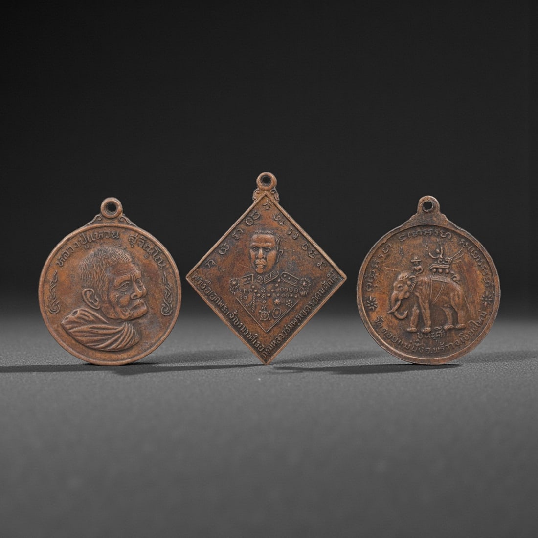 A Group of Three Thai Copper Amulets, 20th Century, 45FL: Each amulet is cast from copper with a warm brown patina, fitted with a suspension loop at the top. The first is a circular medallion depicting a Buddhist monk in profile, with Thai script inscribed a