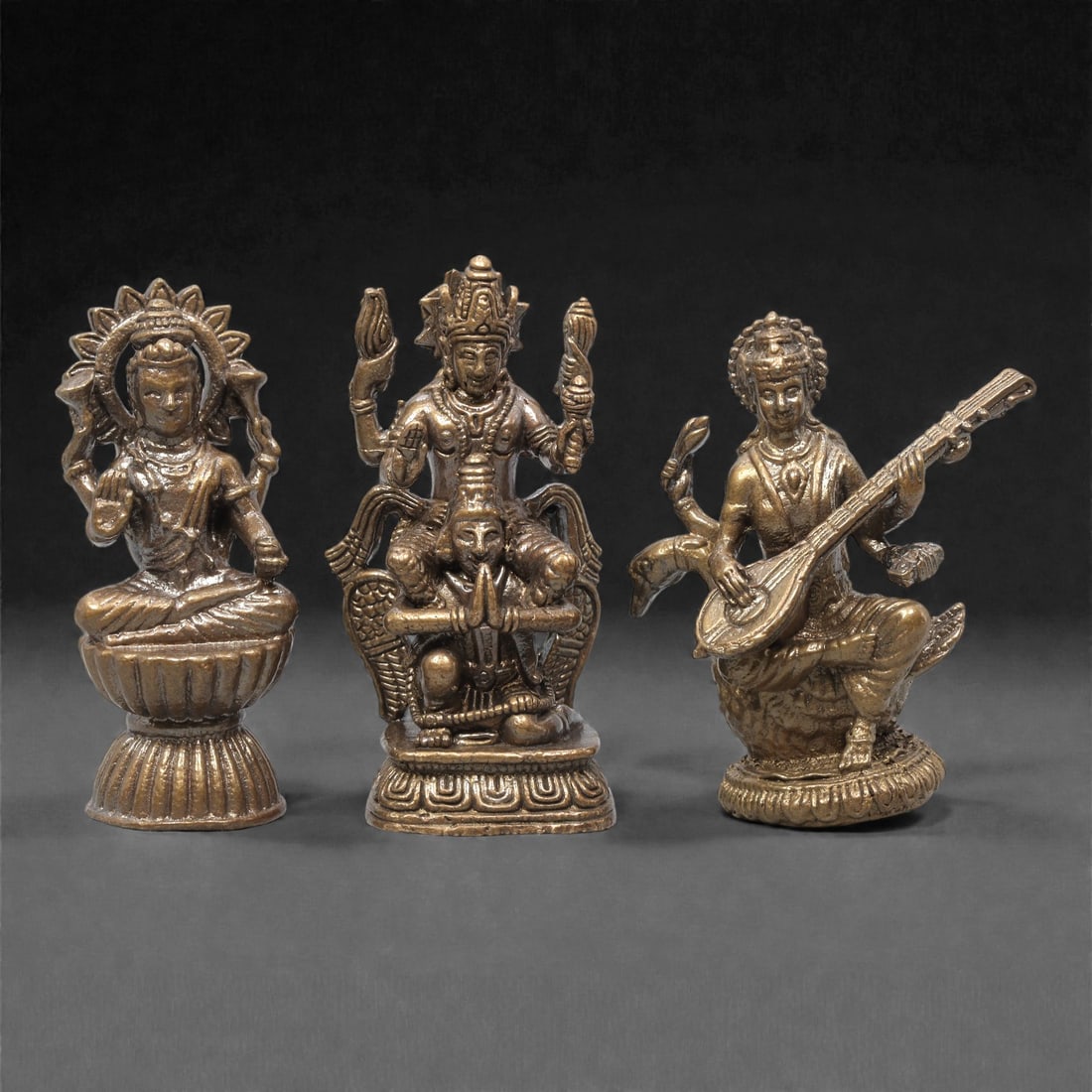 A Set of Three Thai Brass Hindu Deity Figures, 20th Century, 76ZG: Cast from brass with a dark, aged patina, the set comprises three distinct Hindu deity figures. The left figure is seated on a lotus base, with a radiating halo framing its head, multiple arms in symb