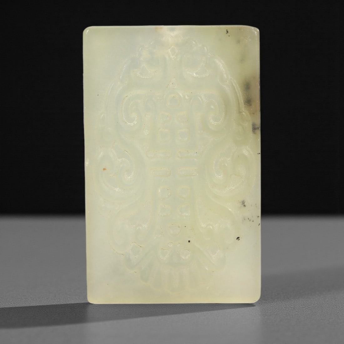 A Chinese Hetian Jade Plaque, 20th Century, 67FH: Carved from translucent, pale celadon Hetian jade with minor dark inclusions, the rectangular plaque features low-relief decoration of symmetrical, voluminous scrollwork flanking a central stylized sh