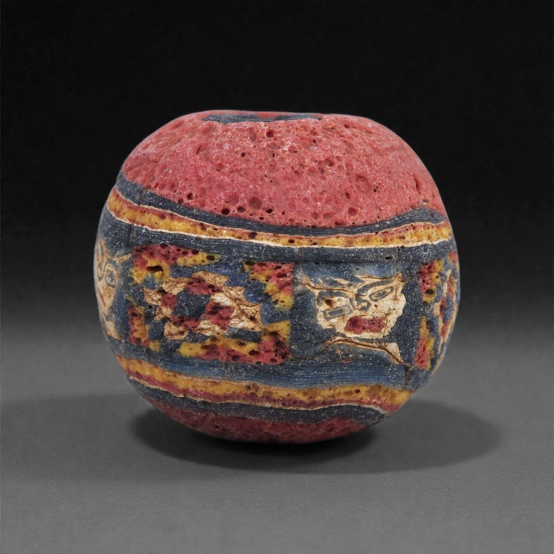 Ancient Glass Bead - Egyptian Face-Mosaic Sphere, 14J: Constructed from multicolored mosaic canes, this bead features a rare anthropomorphic mask motif embedded within a horizontal panel encircling the body. The face, rendered in cream, red, and blue tone