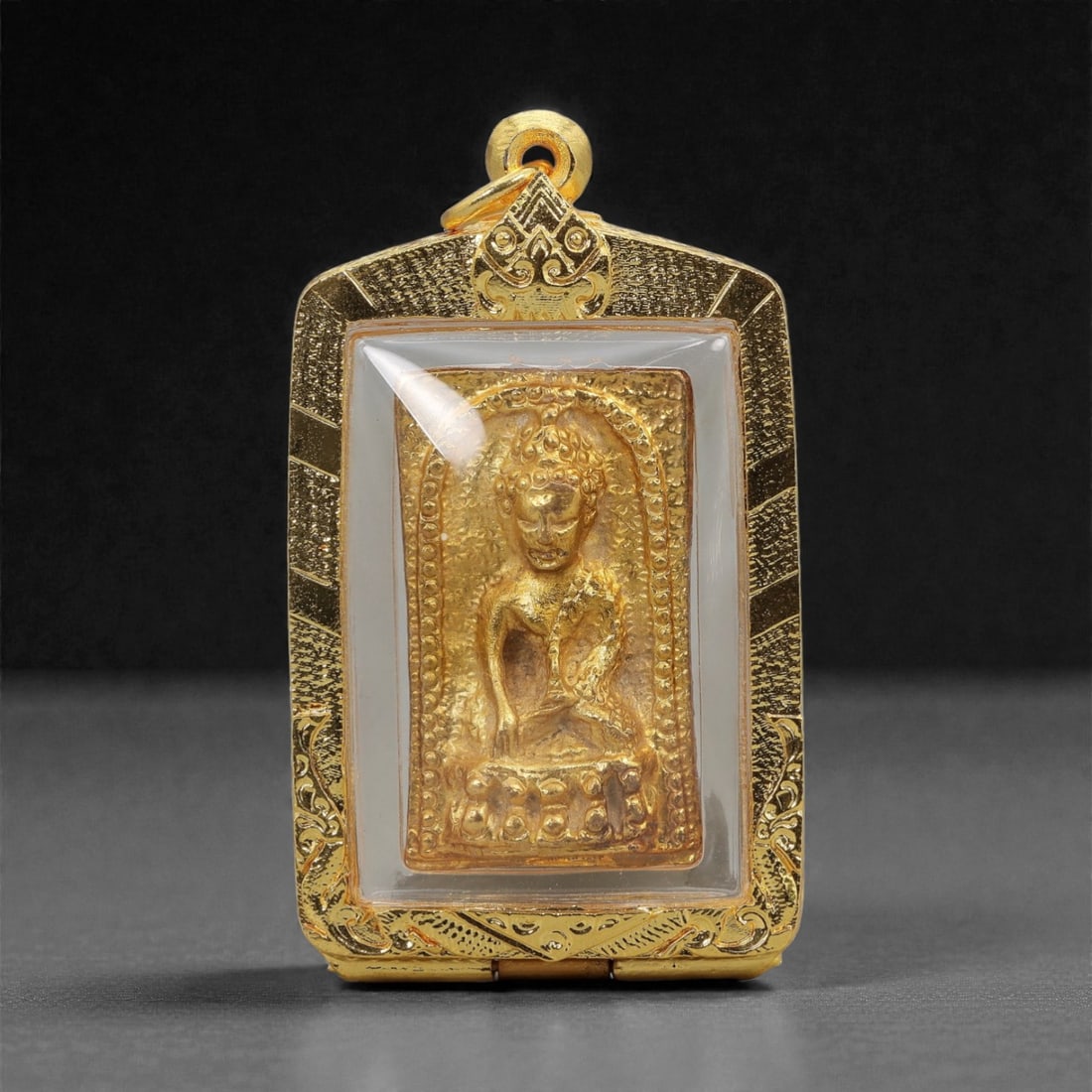 A Thai Buddhist Gilt Metal Amulet in Gilt Metal Frame, 20th Century, 56FN: The amulet is encased in a rectangular gilt metal frame with elaborate chased scrollwork decoration and a suspension loop at the apex. The central plaque is set within a frosted white glass bezel, enc