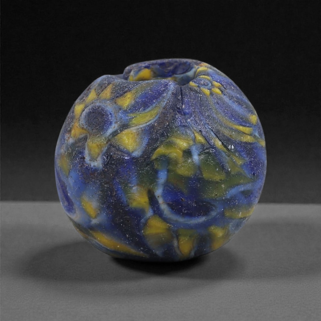 Ancient Glass Bead - Islamic Marvered Polychrome Sphere, 55J: Formed in deep cobalt blue glass enriched with yellow and green trails, this small bead exemplifies early Islamic marvered decoration of the first millennium CE. The molten trails were applied in curv