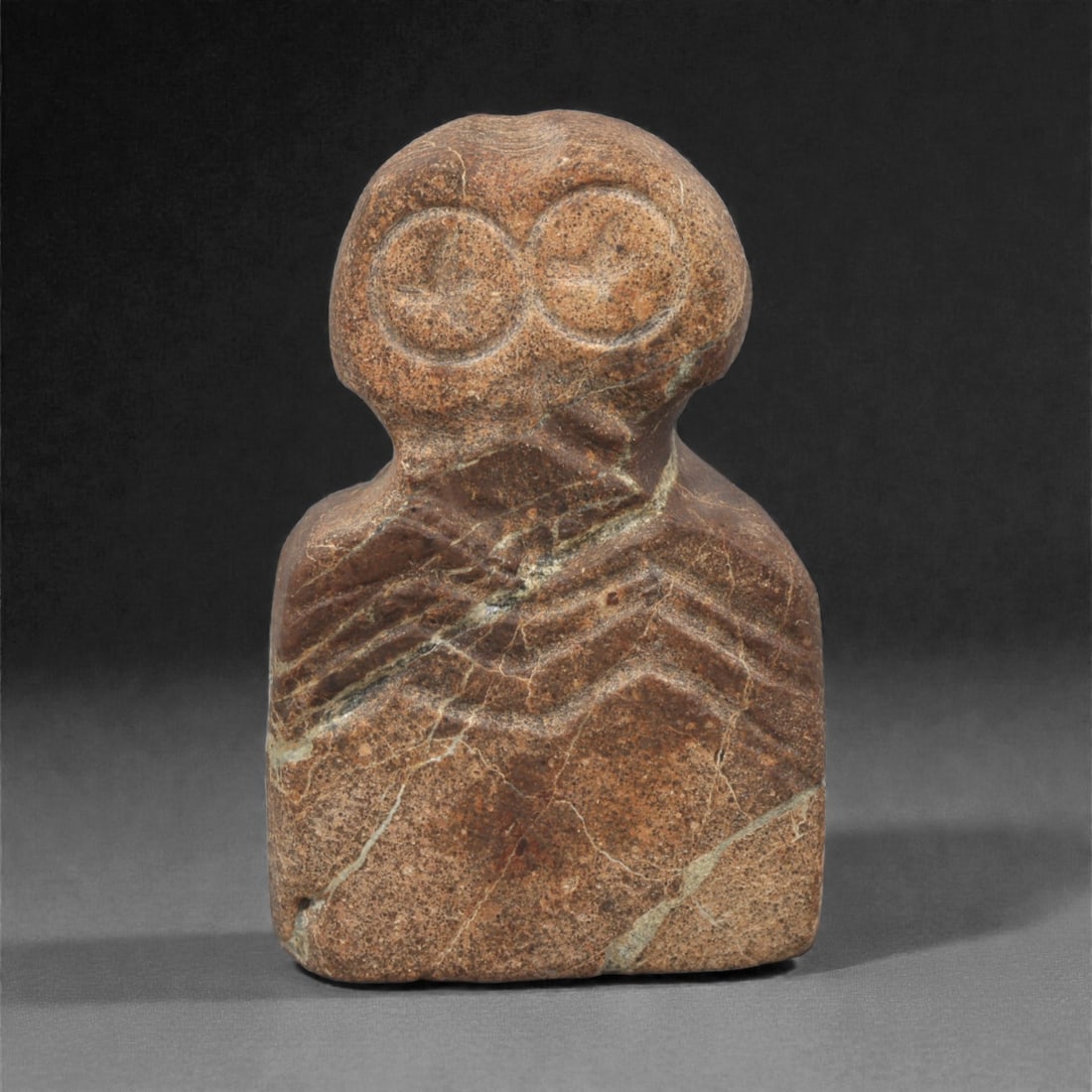 An Ancient Near Eastern Stone Eye Idol, 4th-3rd Millennium BCE, 23AC: Carved from a dense, dark reddish-brown stone with prominent lighter mineral veining and a granular matrix, the idol presents the classic Tell Brak type form: a bulbous, bilobed head surmounting a com