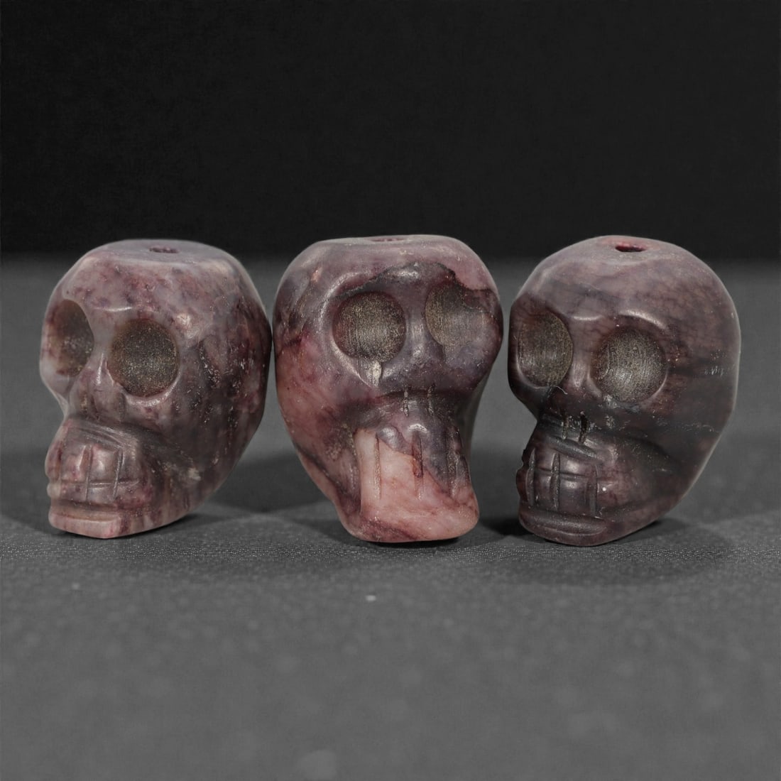 A Group of Tibetan Skull Beads, 20th Century, 43FN: Comprising three beads, each is carved in the form of a human skull, with a central perforation for suspension. The surfaces exhibit natural color variation, ranging from deep purplish-red to dark gre