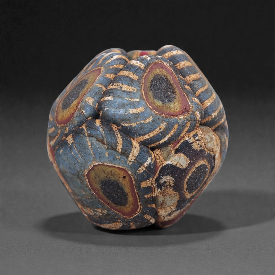 Ancient Glass Bead - Multi-Lobed Roman Eye-Motif Form, 88J: Shaped into a rare multi-lobed configuration, this bead is composed of fused mosaic canes arranged to create concentric eye motifs in blue, yellow, and red tones. The lobed structure results from pres