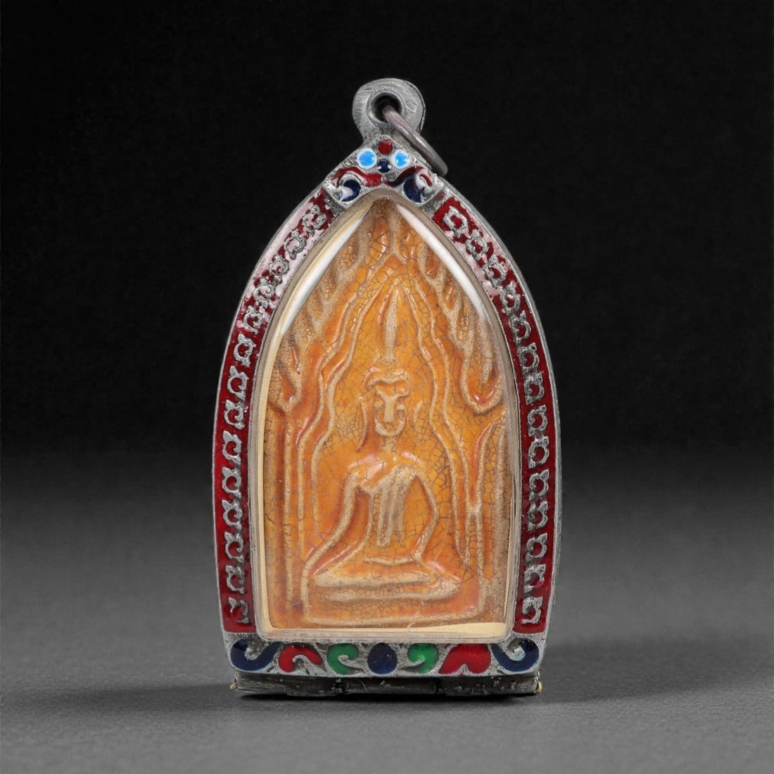 A Thai Buddhist Enameled Metal and Composite Amulet, 20th Century, 46FN: The amulet is housed in a silvered metal frame of arched, tapering form with a suspension loop at the apex. The obverse frame is embellished with applied red, blue, and green enamel inlay within chase