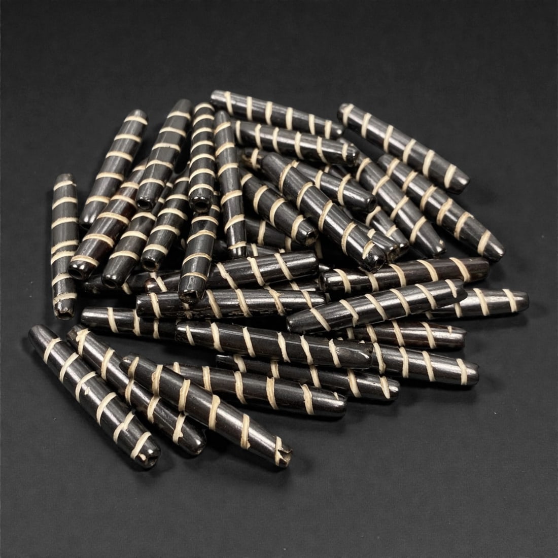 A Lot of Tibetan Bone Beads with Spiral Inlay Motif, 20th Century, 98S: A substantial grouping of elongated, tubular bone beads, characteristic of Tibetan Buddhist devotional art. Each bead is uniformly shaped with a slender, tapered form and features the iconic spiral in