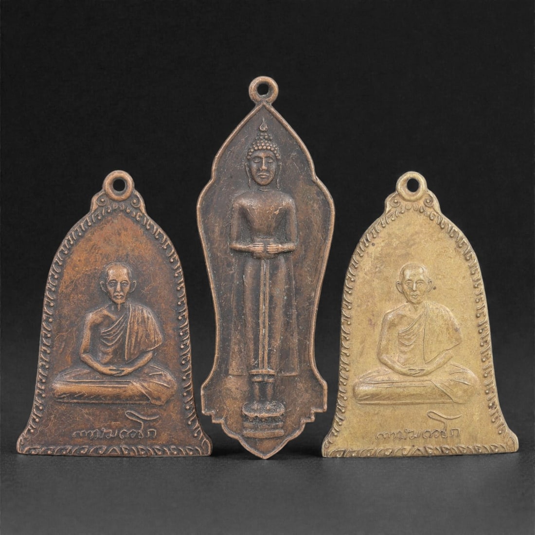 Three Thai Copper Alloy Devotional Amulets, 20th Century, 82FN: Cast from copper alloy with patinas ranging from warm brown to golden brass, the group comprises three distinct devotional amulets, each with a suspension loop at the apex for wear. The first amulet i