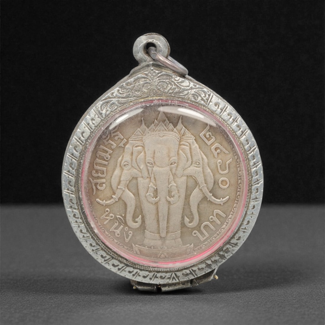 A Thai Silver Coin Amulet in Silver Enamel Case, 20th Century, 89FN: Cast from silver, the amulet is a Thai coin. The obverse depicts a multi-headed elephant (Erawan) with Thai script characters inscribed around the perimeter. The reverse depicts a royal figure in mili