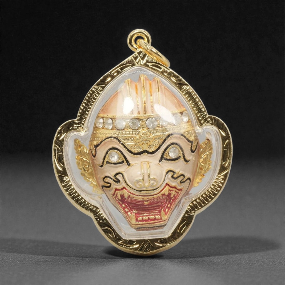 A Thai Hanuman Amulet Pendant, 20th Century, 18FP: This pendant is centered on a painted and gilded Hanuman head, set within a gilded metal frame and encased in a transparent protective cover. The Hanuman figure is rendered with a light pink complexio