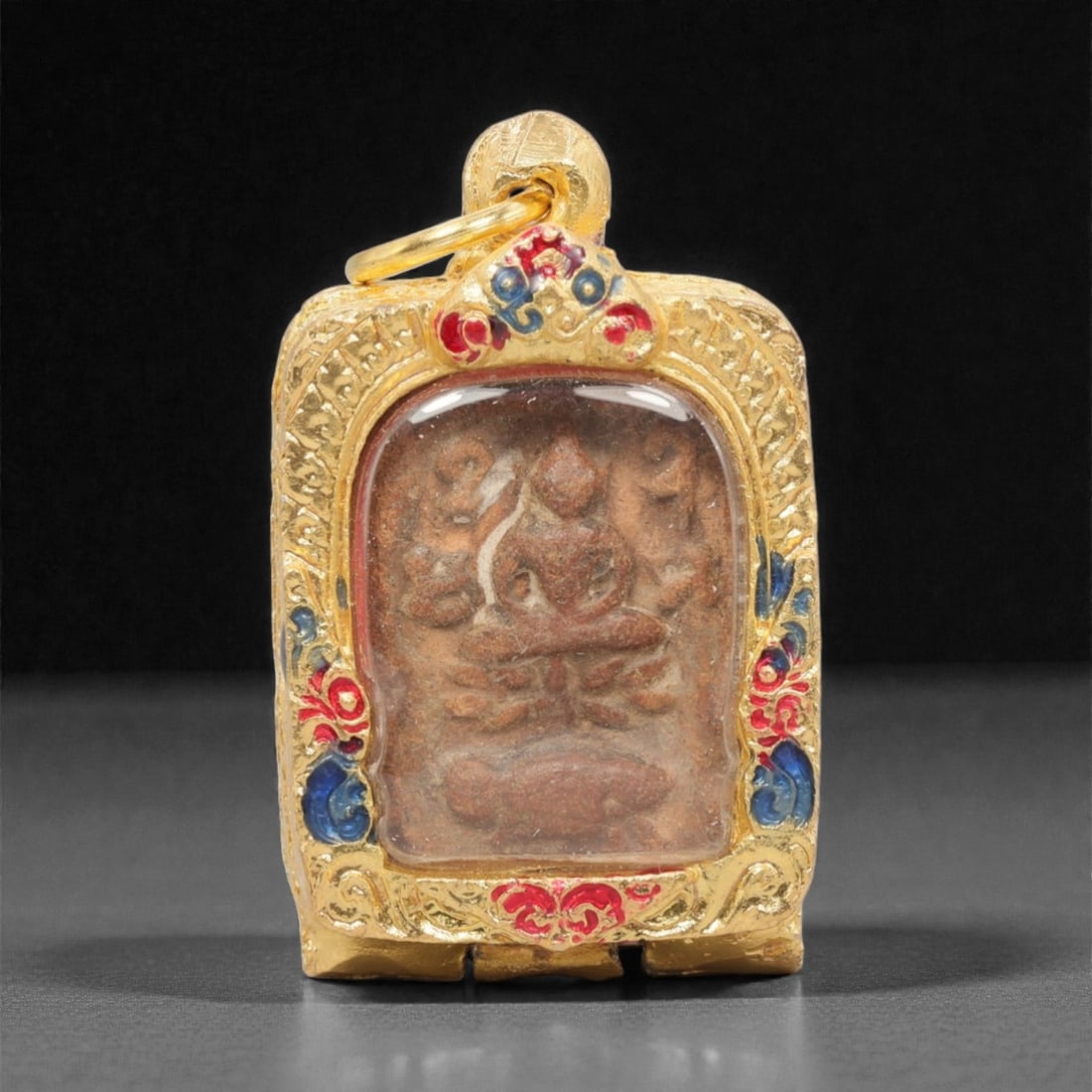 A Thai Clay Buddha Amulet in Gilt Metal Enamel Case, 20th Century, 95FN: Molded from reddish-brown clay with visible surface wear and a distinct linear imperfection, the amulet depicts a seated Buddha in meditative posture, with additional symbolic elements below. The amul
