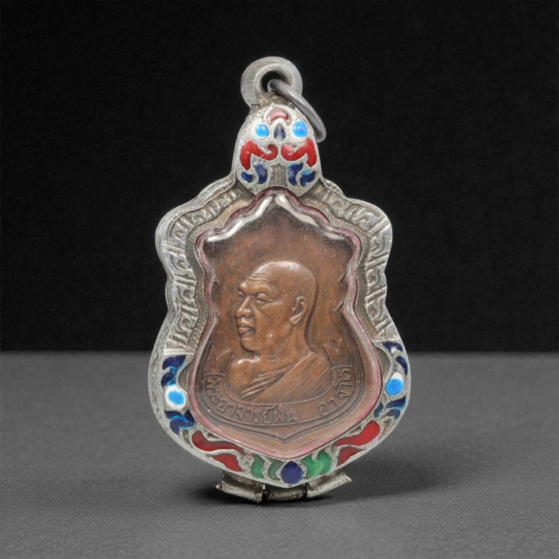 A Thai Monk Amulet Pendant, 20th Century, 17FP: This shield-shaped amulet pendant features a copper core depicting a revered Thai monk, encased in an ornate silver frame with a transparent protective cover, measuring 3.4 x 6 cm (1 5/16 x 2 3/8 in.)