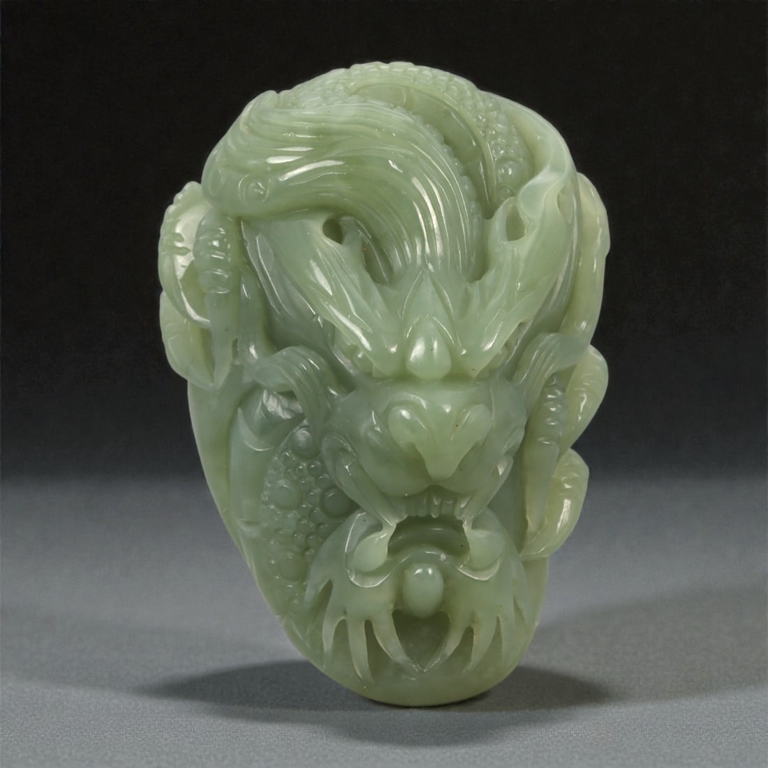 A Chinese Jade Dragon Mask Pendant, 20th Century, 54YA: Carved from pale green jade with soft translucency and subtle tonal variation, the pendant is worked in the round as a stylized dragon mask shown frontally. The face is rendered with prominent arched