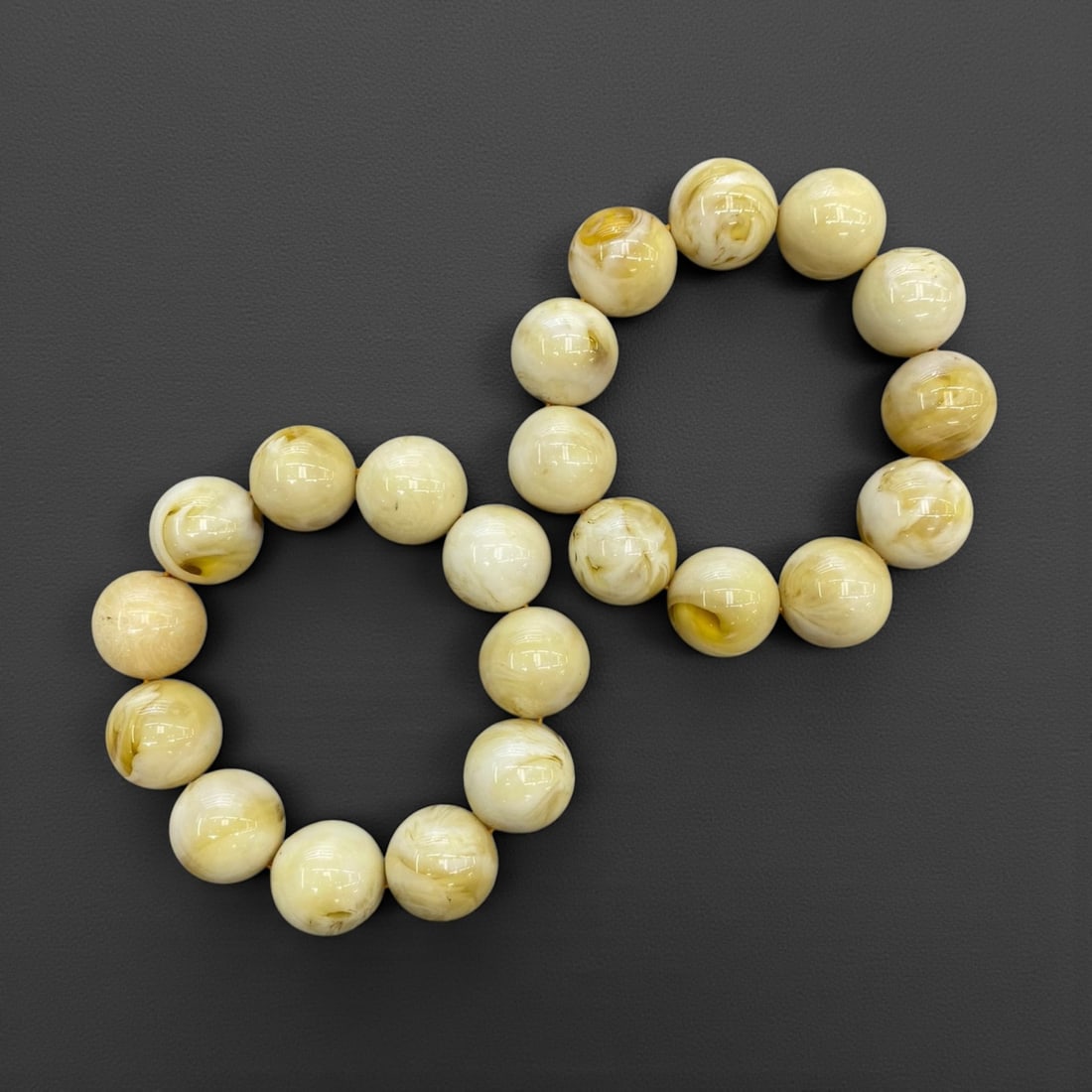 A Group of Two Amber Resin Beaded Bracelets, 149g, 92AA: Comprising two stretch bracelets composed of polished spherical amber resin beads. The beads exhibit a creamy, off-white base with subtle golden-yellow marbling and translucency, characteristic of the