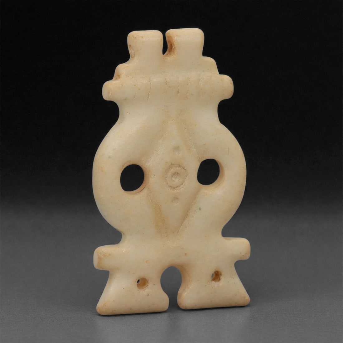 A Sino-Mongolian Jade Amulet, Medieval Period, 10th-14th Century CE, 25LQ: Carved from pale cream-colored jade with a smooth, slightly lustrous surface, the amulet is shaped as a vertically oriented openwork form with a bifurcated upper terminal pierced by two small perforat