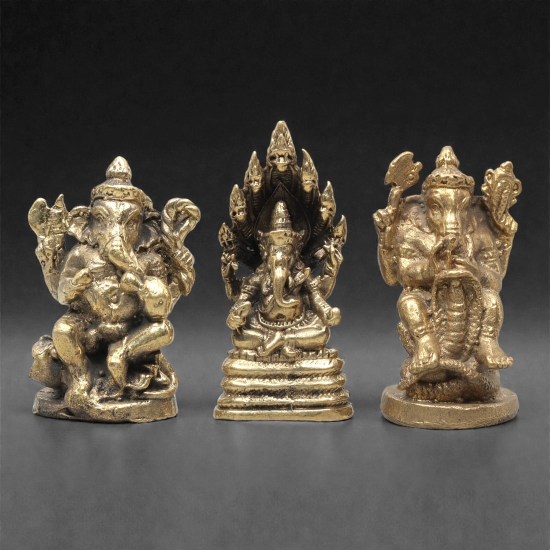 A Set of Three Thai Brass Ganesha Figures, 20th Century, 63ZG: Cast from brass with a warm golden patina, the set comprises three distinct Ganesha figures. The left figure is seated, with multiple arms holding symbolic attributes, and a rat mount at its feet. The