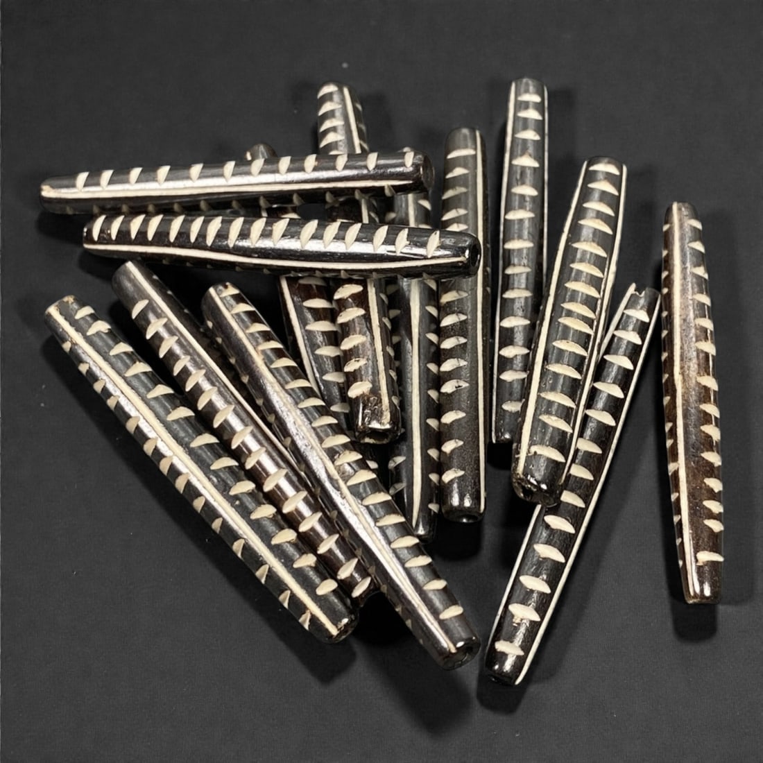 A Lot of Tibetan Bone Beads with Chevron Inlay Motif, 20th Century, 82S: A substantial grouping of elongated, tubular bone beads, characteristic of Tibetan Buddhist devotional art. Each bead is uniformly shaped with a slender, tapered form and features the iconic chevron i