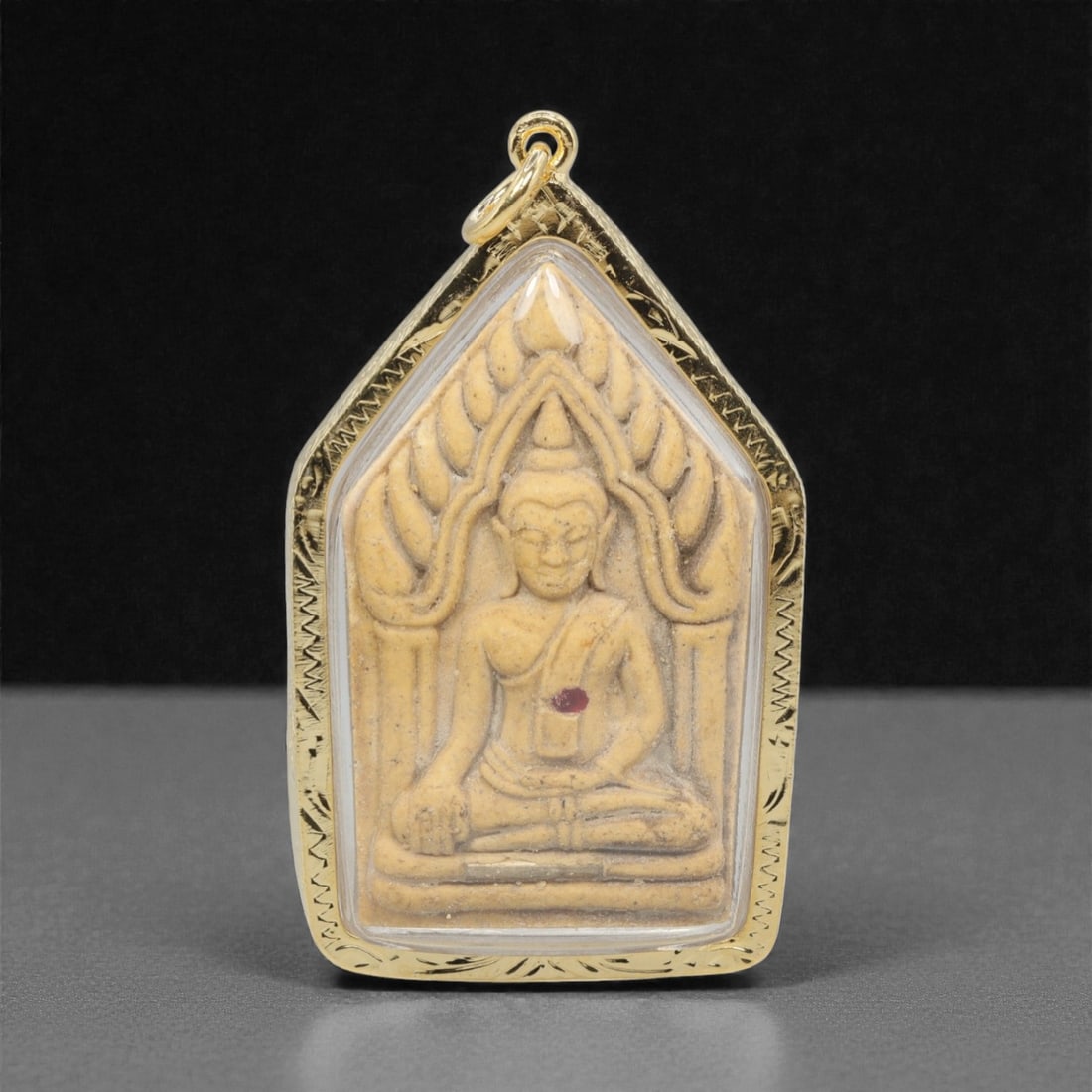 A Thai Khmer-Style Buddha Amulet Pendant, 20th Century, 27FP: This pendant features a light beige composite amulet core, encased in an ornate gilded metal frame with a transparent protective cover, measuring 6.3 x 3.6 cm (2 1/2 x 1 7/16 in.). The obverse display