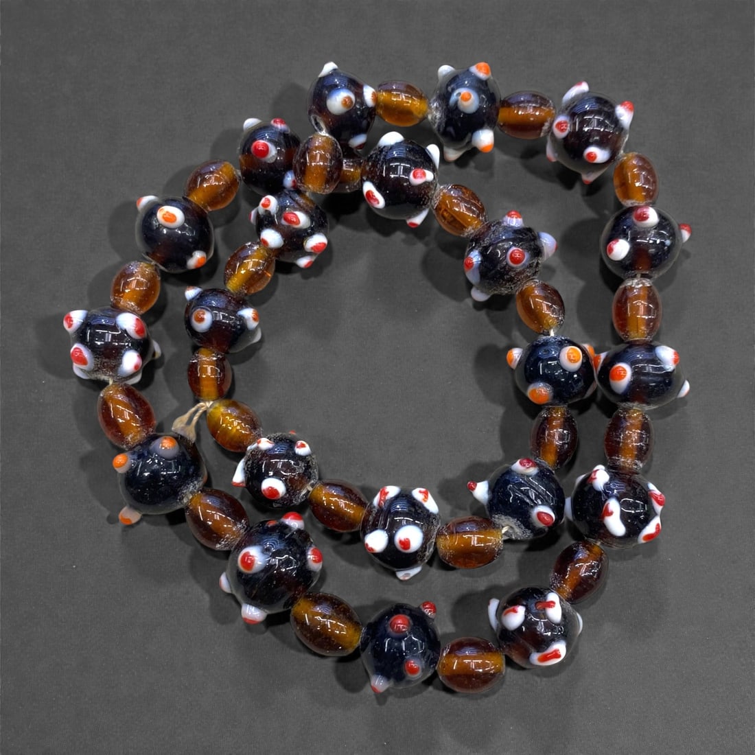 A Mid 20th Century Glass Bead Necklace Strand, 148g, 94AA: Composed of two distinct types of glass beads strung in an alternating pattern. The primary beads are spherical, with a dark translucent base, decorated with raised white and red eye-like motifs, each