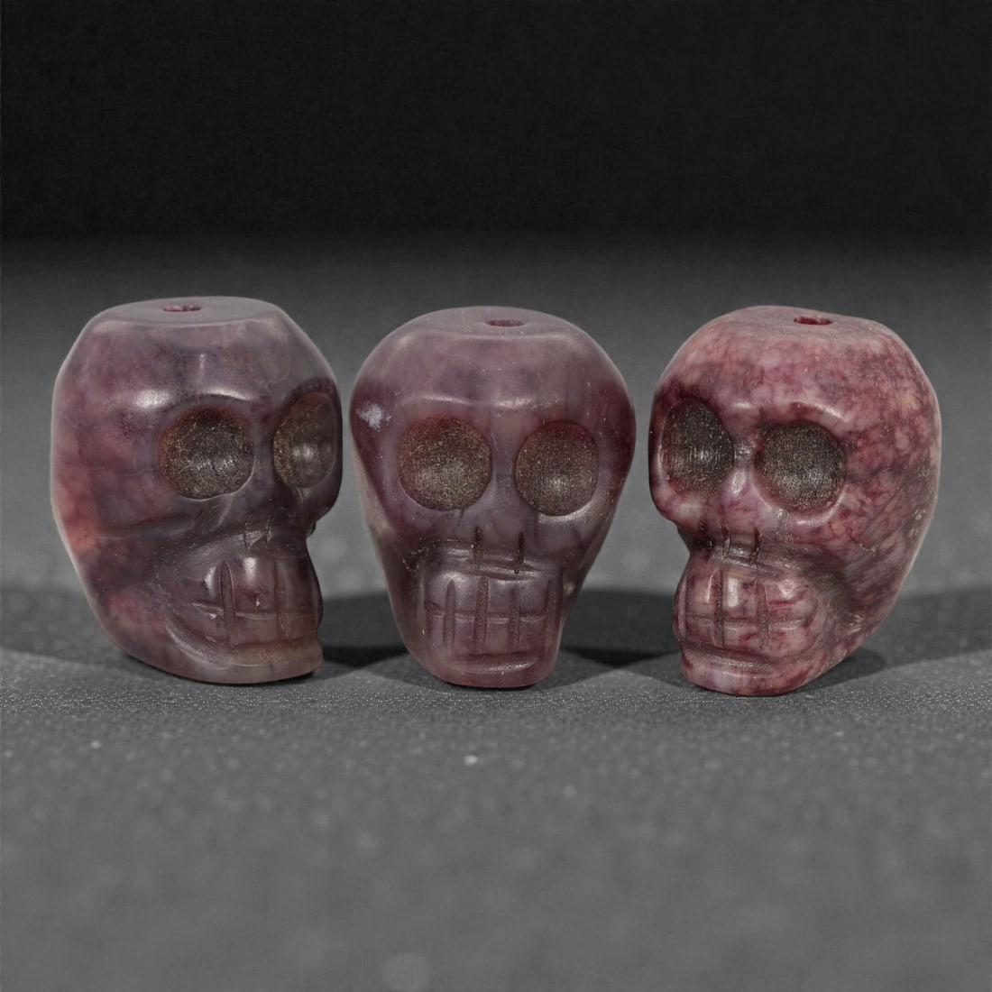 A Group of Tibetan Skull Beads, 20th Century, 44FN: Comprising three beads, each is carved in the form of a human skull, with a central perforation for suspension. The surfaces exhibit natural color variation, ranging from deep purplish-red to marbled 