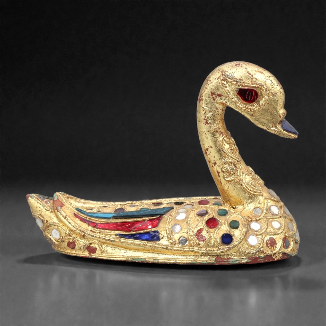 A Southeast Asian Gilt Wood Swan Figure, 20th Century, 51FK: Carved from wood and modeled as a stylized swan shown resting with an arched neck and forward-facing head, the figure is decorated overall with gilt surface treatment and inset colored glass elements.