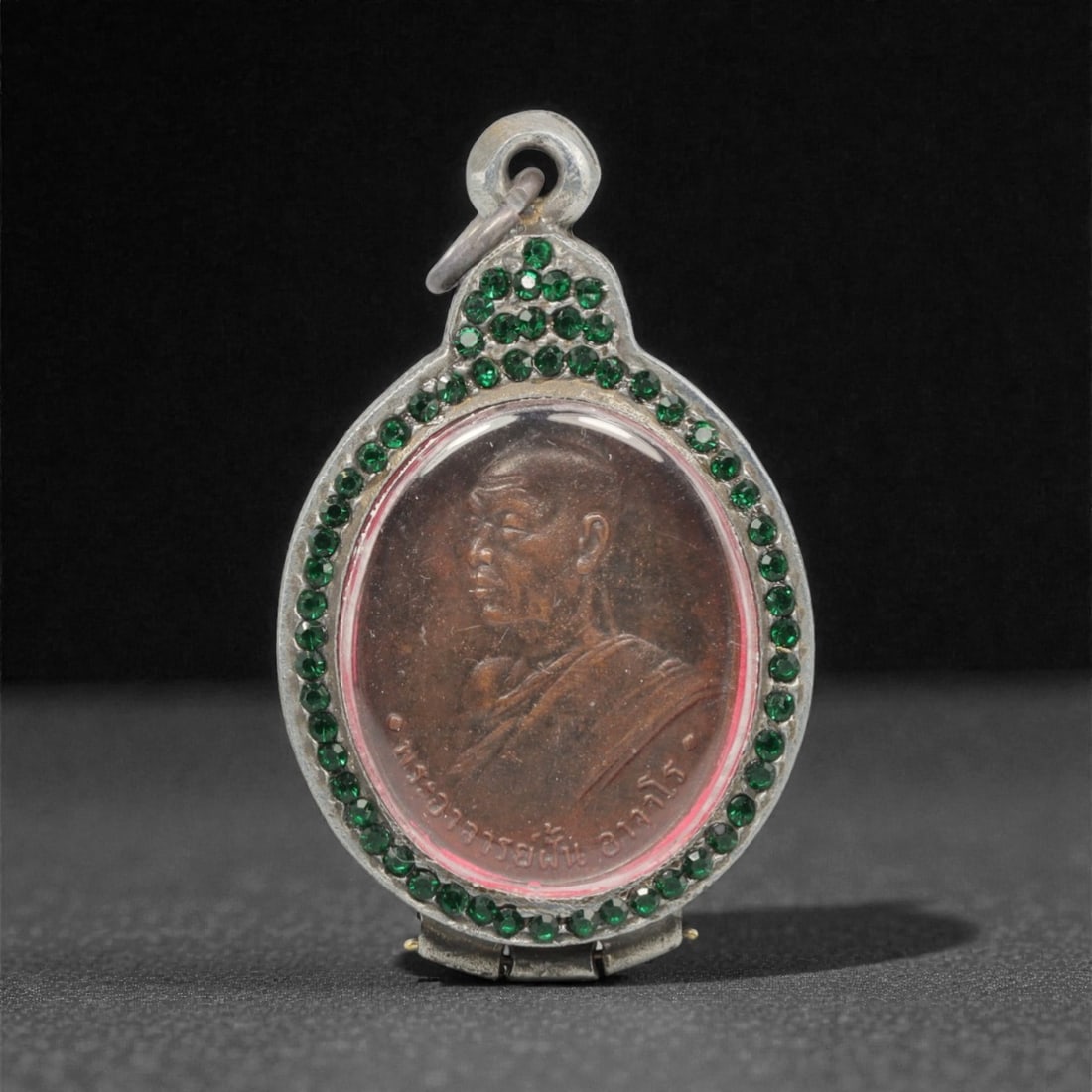 A Thai Monk Amulet Pendant with Green Paste-Set Frame, 20th Century, 14FP: This oval amulet pendant features a patinated copper alloy core, encased in an ornate silver frame with a transparent protective cover, measuring 5.7 x 3.5 cm (2 1/4 x 1 3/8 in.). The obverse displays
