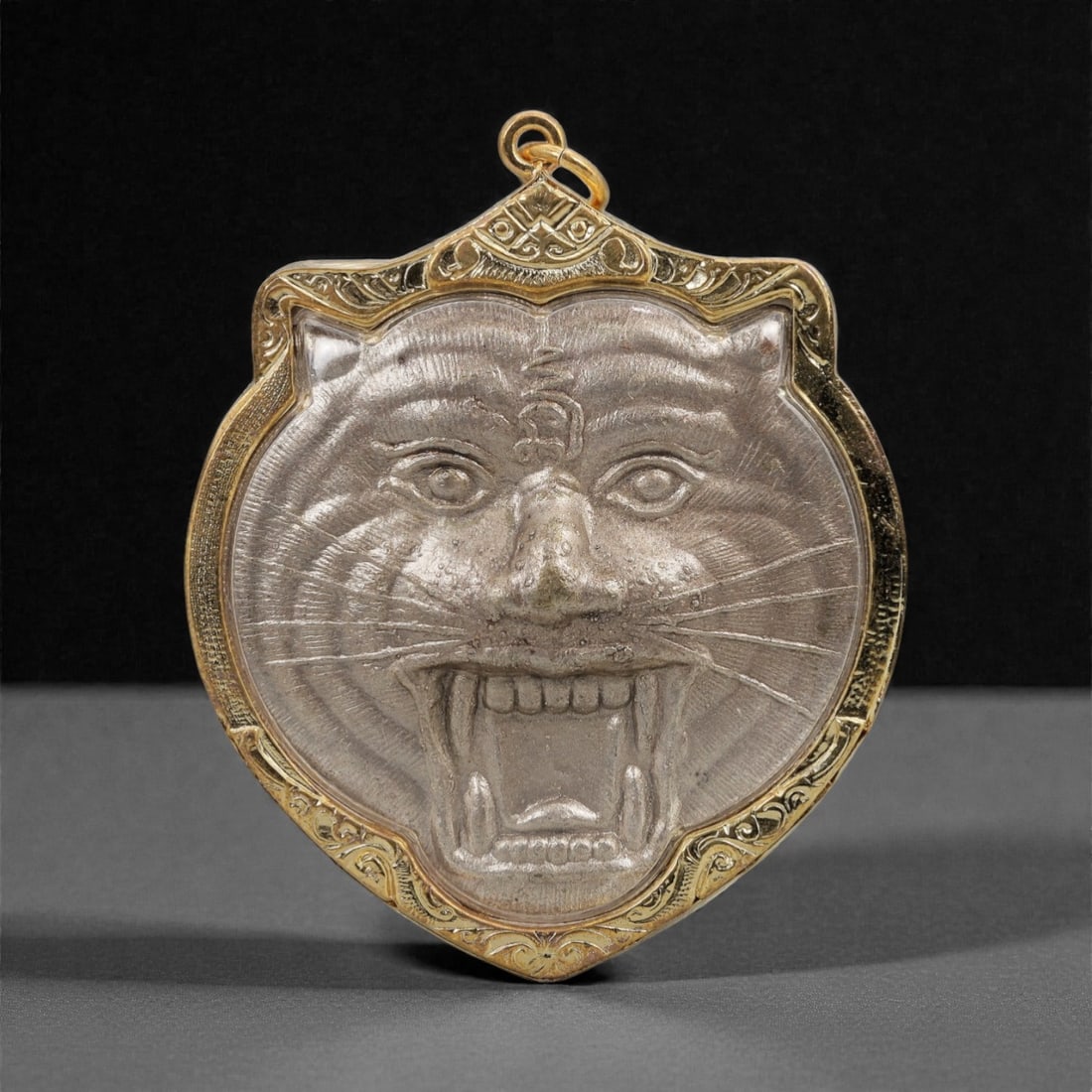 A Thai Silvered Metal Amulet in Gilt Metal Frame, 20th Century, 61FN: The amulet is encased in a shield-shaped gilt metal frame with incised scrollwork borders and a suspension loop at the apex. The obverse features a silvered metal relief of a seated Buddhist monk in m