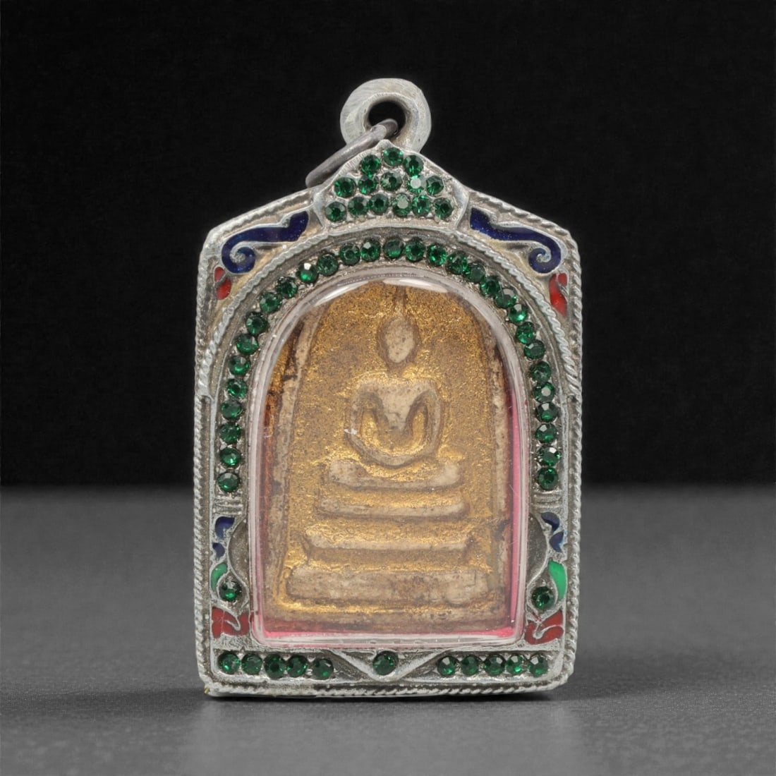 A Thai Clay Buddha Amulet in Silver Enamel and Gem-Set Case, 20th Century, 96FN: Molded from light tan clay with surface gilding and patination, the amulet depicts a seated Buddha in meditative posture atop a multi-tiered base. The reverse is heavily patinated, with a distinct hea