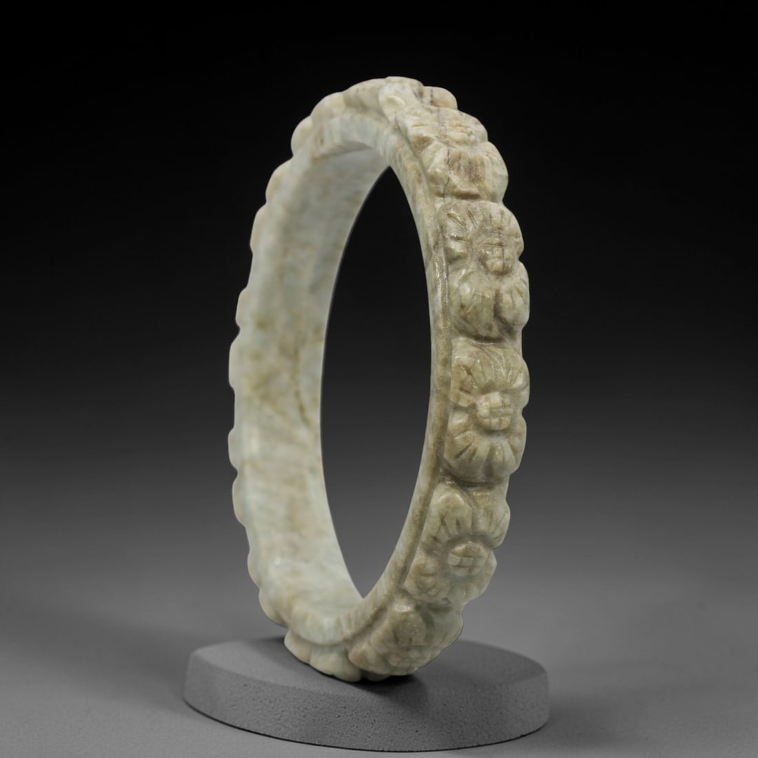 A Chinese Carved Jadeite Jade Bangle Bracelet, 20th Century, 77ZL: A fine jadeite bangle of flattened circular form, masterfully carved in relief with a continuous band of chrysanthemum blossoms. The stone exhibits a pale milky-green base, accented by subtle brownish