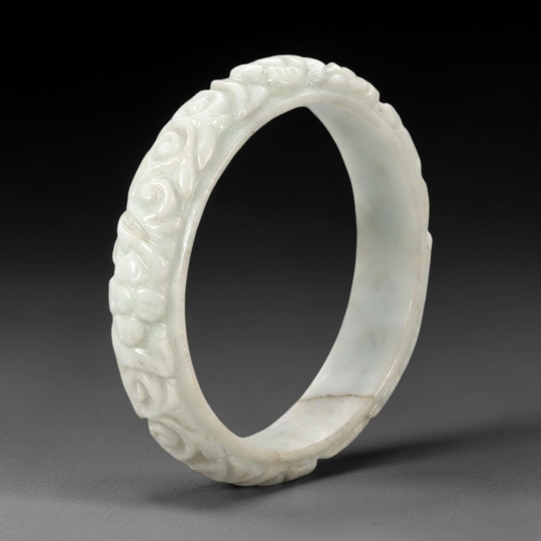 A Chinese Carved Jadeite Jade Bangle Bracelet, 20th Century, 32ZL: A fine jadeite bangle of flattened circular form, masterfully carved in relief with a continuous band of scrolling foliate and floral motifs. The stone exhibits a translucent pale white base, with sub