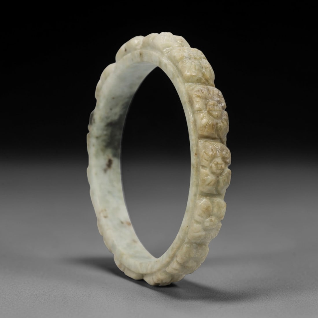 A Chinese Carved Jadeite Jade Bangle Bracelet, 20th Century, 80ZL: A fine jadeite bangle of flattened circular form, masterfully carved in relief with a continuous band of chrysanthemum blossoms. The stone exhibits a pale milky-green base, accented by subtle brownish