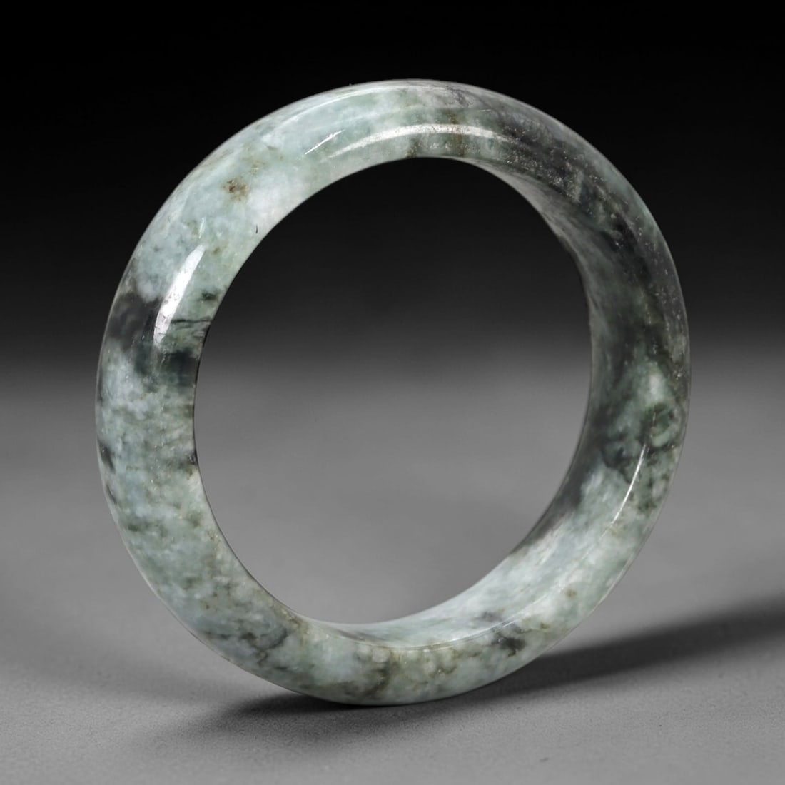 A Chinese Jadeite Jade Bangle Bracelet, 20th Century, 89ZK: Carved from jadeite jade with a pale green base and irregular dark grey-green mottling, the bangle is formed as a continuous circular band with a rounded cross-section. The surface is smoothly polishe