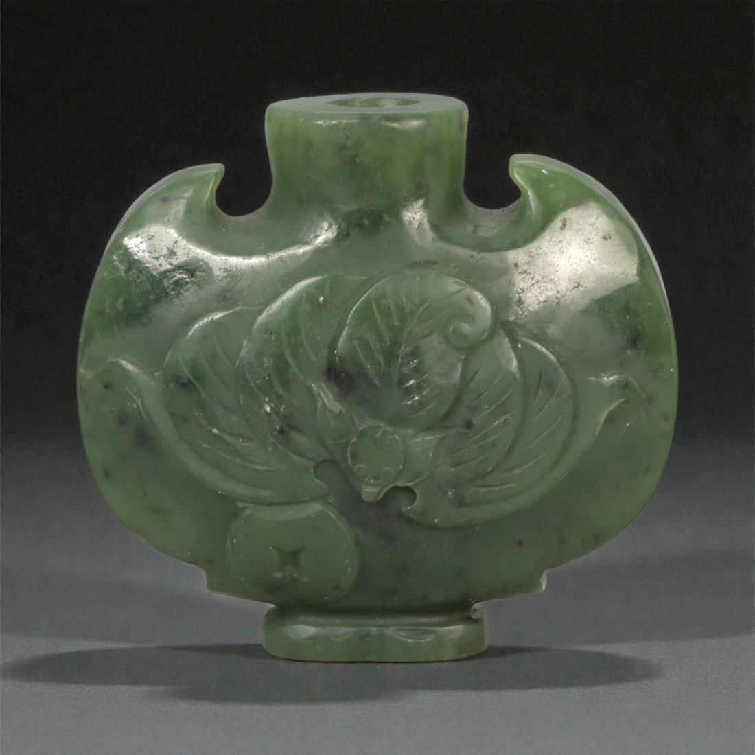 A Chinese Jade Lotus-Decorated Snuff Bottle, 20th Century, 19YA: Carved from dark green jade with lighter translucent areas and scattered natural inclusions, the vessel is formed as a flattened moonflask-shaped snuff bottle with a short cylindrical neck and raised