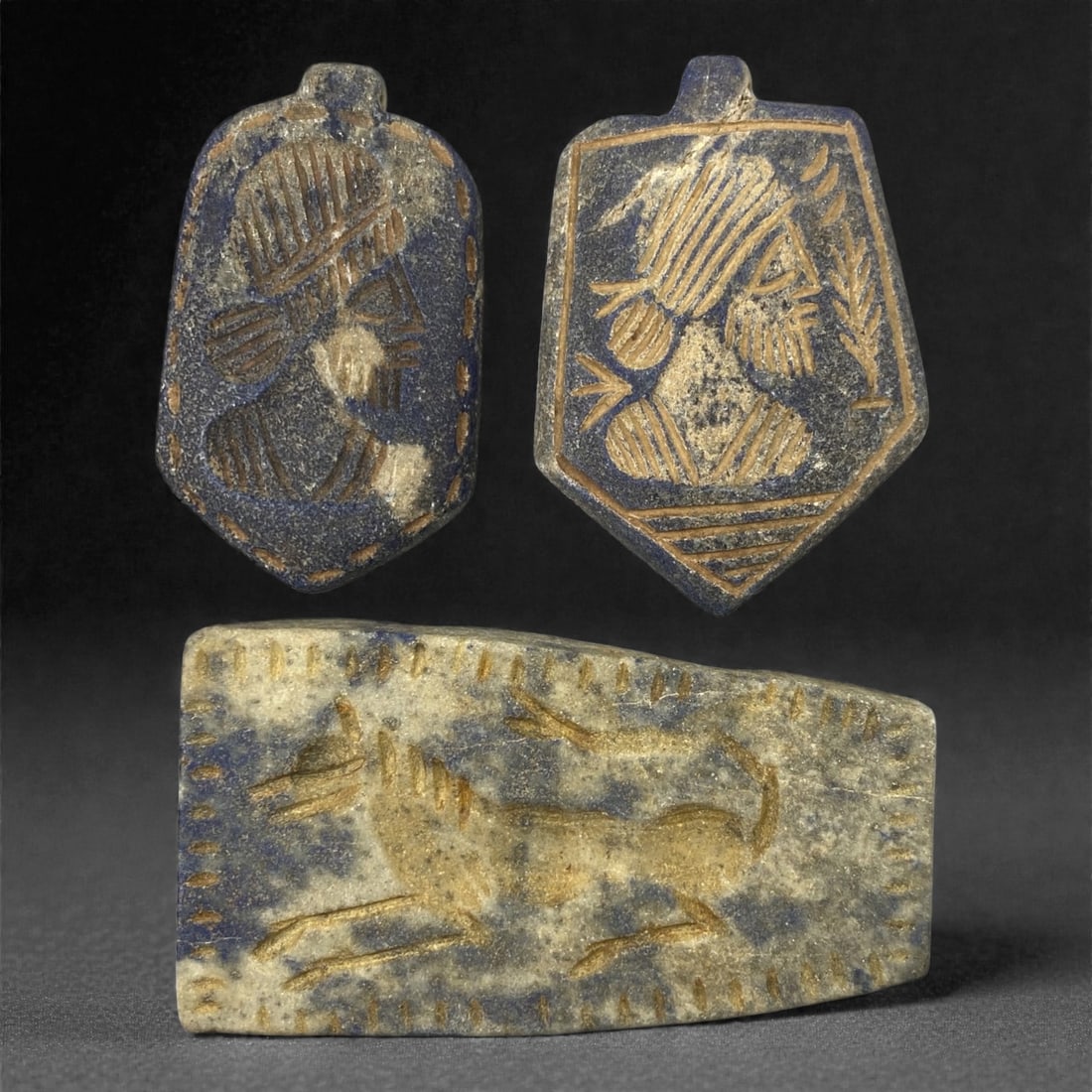 A Group of Three Sasanian Lapis Lazuli Plaque and Amulets, 3rd-7th Century CE, 54FM: Comprising three lapis lazuli carvings, each with a deep blue matrix and visible calcite inclusions, exhibiting natural surface patina and wear. The top-left shield-shaped amulet depicts a profile bus