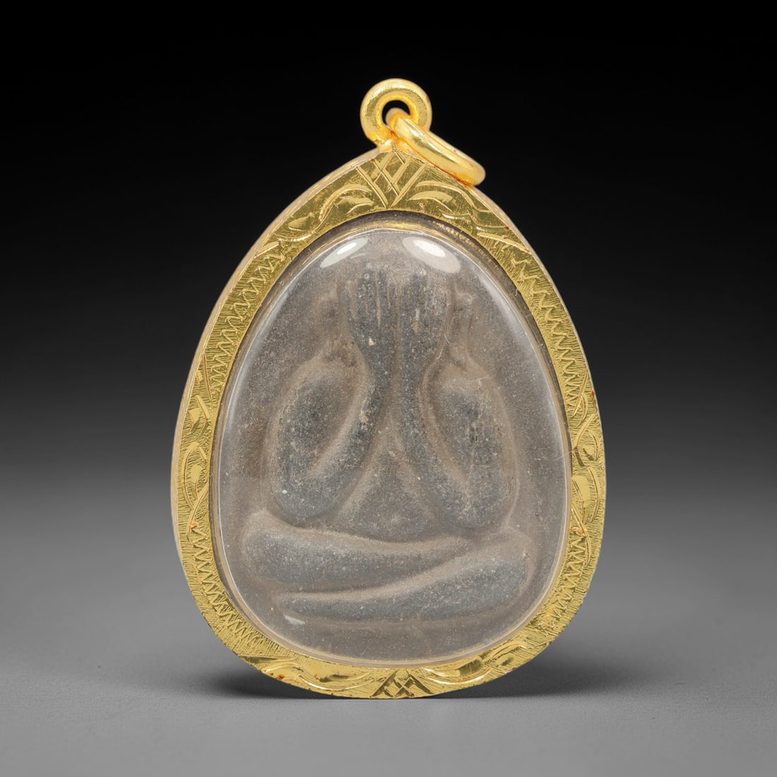 A Thai Phra Pidta Amulet in Gold Frame, Late 20th Century, 2FR: A teardrop-shaped amulet, measuring 4.8 x 3.3 cm, set within an ornate gold frame with a suspension loop. The obverse depicts the Buddha Phra Pidta in seated meditative posture, with hands covering th