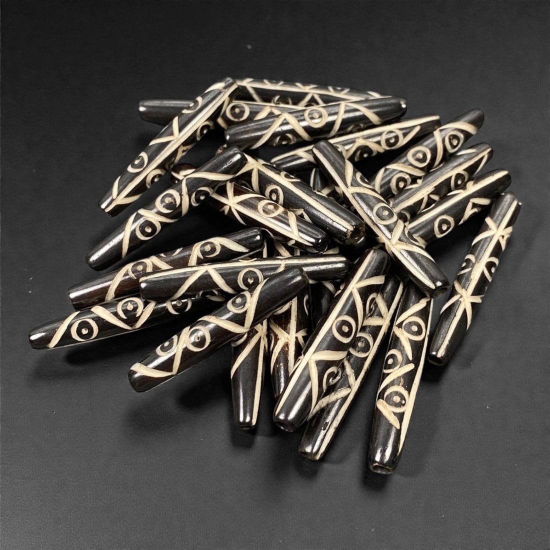 A Tibetan Bone Bead Group, Inlaid Scroll and Linear Pattern, 20th Century, 75S: Composed of animal bone, each bead features an elongated cylindrical form with a tapered end, consistent in proportions across the group. The surface displays an inlaid decorative scheme combining scr