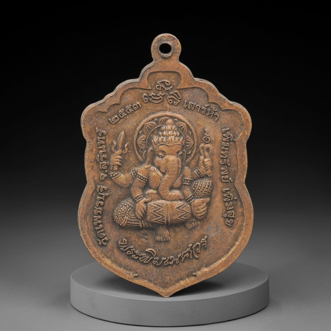 A Thai Copper Alloy Ganesha Amulet, Late 20th Century, 96FP: A shield-shaped copper alloy amulet, measuring 4.5 x 2.7 cm. The obverse depicts the four-armed deity Ganesha in seated meditative posture, holding a goad and a noose, with Thai script inscriptions en