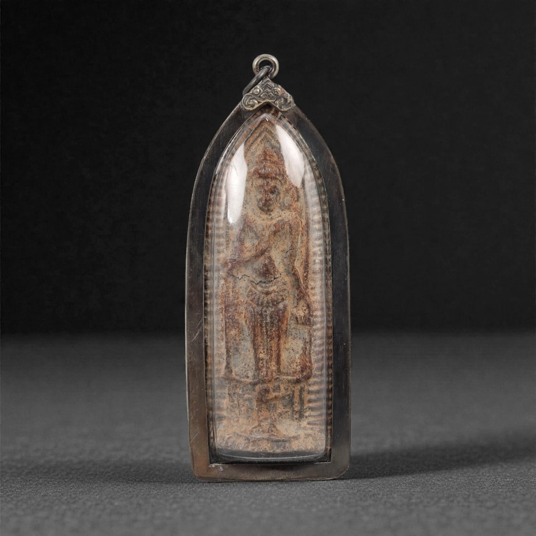 A Thai Standing Buddha Amulet with Silvered Frame, 20th Century, 37FP: The amulet is of elongated, teardrop-shaped form, featuring a molded standing Buddha figure on the obverse, rendered in a dark patinated material against a light beige ground. The Buddha is depicted w
