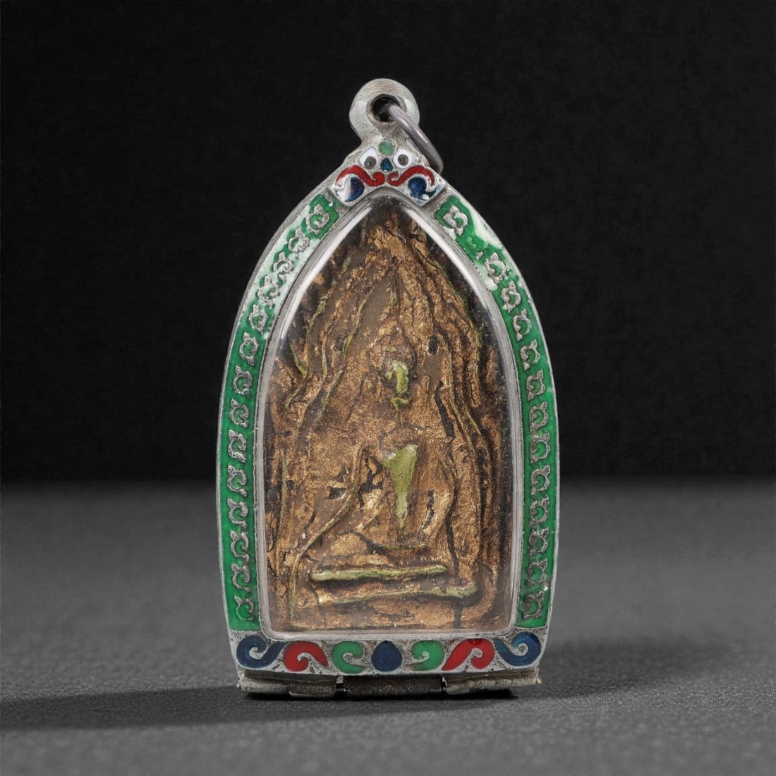 A Thai Gilded Clay Buddha Amulet in Silver and Enamel Case, 20th Century, 46ZH: The amulet is formed from gilded clay, depicting a seated Buddha in bhumisparsha mudra, with the right hand touching the earth and the left hand resting in the lap in dhyana mudra. The figure is set w