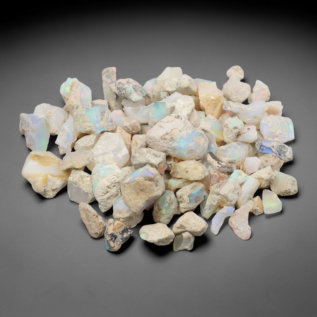 An Ethiopian Welo Opal Rough Lot, 50.90g, 47ZH: A parcel of rough Welo opal, with individual pieces ranging from 1-2 cm in size. The specimens exhibit a milky to translucent base, with visible play-of-color displaying flashes of blue, green, yellow