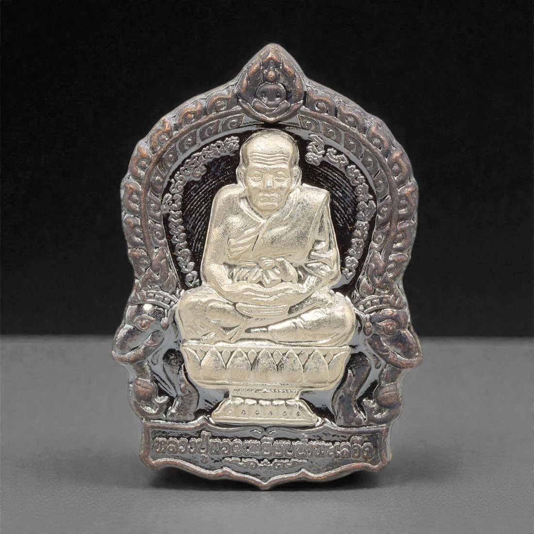 A Thai Bi-Metallic Monk Portrait Amulet, 21st Century, 80FP: Cast with a bi-metallic construction, the amulet has a shield-shaped form with a pointed upper apex. The obverse features a high-relief silver-toned bust portrait of a seated Buddhist monk, set agains