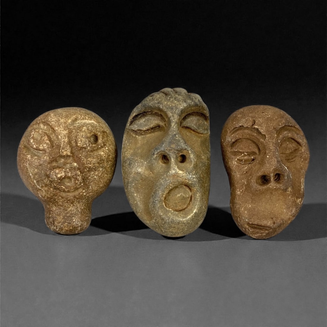 A Group of Three Medieval Stone Maskette Amulets, 10th-14th Century, 49FP: The group comprises three stone maskette amulets, each carved from distinct stone types with varied surface treatments. The first is a brown stone mask with a rounded form and short neck, featuring in